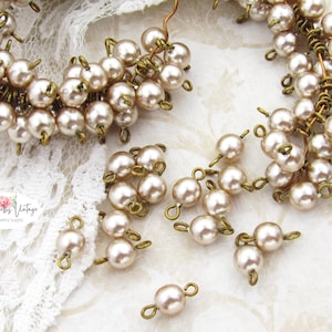 Puede incluir: Primer plano de componentes de joyería de estilo vintage. La imagen muestra grupos de perlas de imitación de color crema, conectadas con hallazgos de metal con tonos de latón antiguo. Algunos componentes están dispuestos en forma circular, otros sueltos.