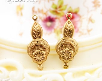 Puede incluir: Dos abalorios de pendientes de metal dorado con diseño floral. Cada abalorio presenta un centro redondo con una flor y un diseño de hoja encima.