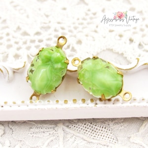 May include: Two green glass oval-shaped cabochons with gold metal settings and loops. The cabochons have a raised design and are set in a gold metal frame. The loops are attached to the frame and can be used to attach the cabochons to a necklace or bracelet.