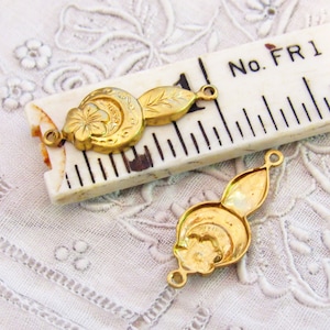 May include: Two gold-toned metal charms with a floral design and a loop on each end. The charms are on a white surface with a ruler in the background. The ruler has the text "No. FR1 2".