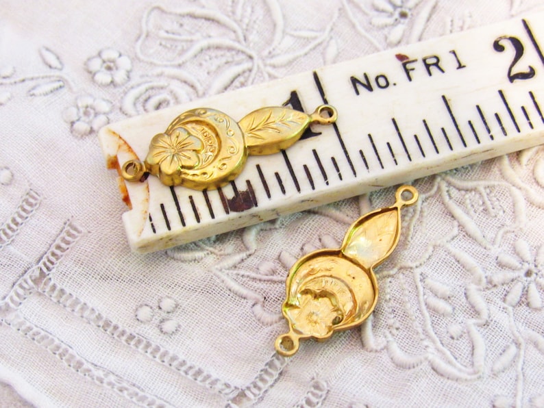 May include: Two gold-toned metal charms with a floral design and a loop on each end. The charms are on a white surface with a ruler in the background. The ruler has the text "No. FR1 2".