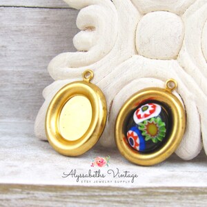 May include: Two gold-toned metal pendant settings, one with a clear glass insert and the other with a colorful Murano glass insert. The settings have a rounded oval shape with a raised edge.