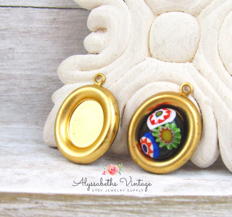 May include: Two gold-toned metal pendant settings, one with a clear glass insert and the other with a colorful Murano glass insert. The settings have a rounded oval shape with a raised edge.