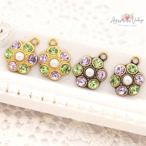 May include: Four vintage-style flower charms with a gold-tone finish. Each charm features a central pearl or white bead surrounded by alternating green and lavender-colored crystals. The charms are designed for jewelry making.