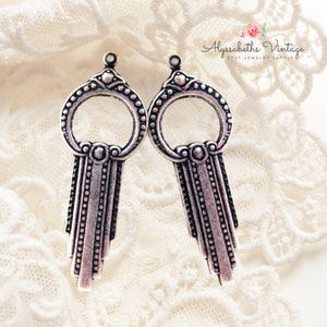 Peut inclure: Une paire de boucles d'oreilles en argent en forme de clé avec un motif circulaire et une frange perlée. Les boucles d'oreilles sont sur un fond de dentelle blanche.