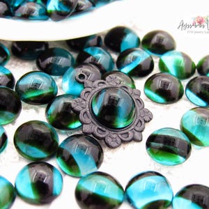 May include: A collection of round, translucent teal and black glass cabochons scattered alongside a single dark metal pendant setting featuring a decorative scalloped edge and a central circular opening for a cabochon insert.