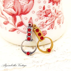 May include: Two vintage-style charms, one silver-toned and one gold-toned, each featuring a rectangular design with three red crystal accents and a circular ring. The charms are set against a white background with a floral pattern.