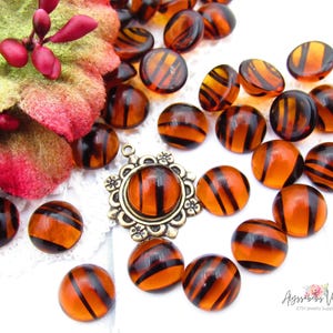 May include: A collection of round, translucent amber-colored glass cabochons featuring dark brown tiger-stripe patterns, accompanied by a single antique bronze-toned metal pendant setting with a floral border and a small red fabric flower.