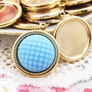 May include: Two gold-toned metal pendant trays with a round shape. One tray has a light blue glass cabochon with a textured surface. The other tray is empty.