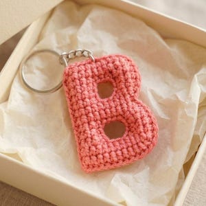 Puede incluir: Un llavero con la letra B tejida a ganchillo en color coral, que cuenta con una anilla de metal plateado en la parte superior. La letra está elaborada con un patrón de punto texturizado y se presenta dentro de una caja de cartón abierta.