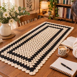 Op de afbeelding: Een rechthoekige gehaakte tafelloper met een herhalend granny square patroon in crème en zwart garen. De loper heeft een geschulpte rand in crème en ligt in het midden op een houten tafel, met een geometrisch ontwerp in afwisselende kleuren.