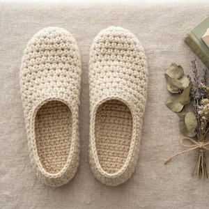 Può includere: Un paio di pantofole all'uncinetto color crema caratterizzate da un motivo a punti testurizzati sull'esterno e una suola interna piatta e solida. Le pantofole sono posizionate fianco a fianco, con una punta arrotondata e una forma aperta.