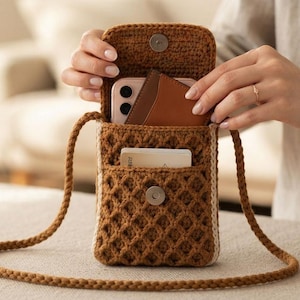 Può includere: Una borsa a tracolla in uncinetto marrone con motivo a rombi, chiusura magnetica e una lunga tracolla intrecciata. La borsa contiene uno smartphone di colore chiaro e un portacarte in pelle marrone, con una carta bianca nella tasca.
