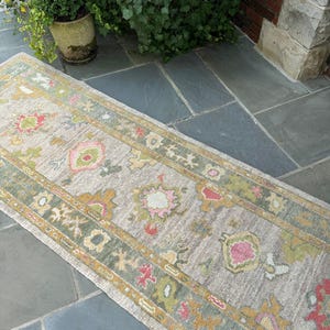 May include: A long, rectangular runner rug featuring a light beige base with a repeating pattern of floral and geometric motifs in muted shades of sage green, mustard yellow, and soft pink, finished with a decorative border along the edges.