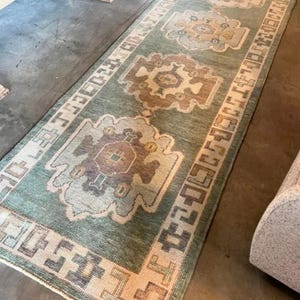 May include: A long rectangular runner rug featuring a sage green background with three repeating geometric medallions in shades of brown, tan, and cream. The rug is framed by a wide border with a repeating block pattern in muted earth tones.