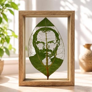 May include: A rectangular light-toned wooden frame containing a single green leaf. The leaf features a detailed, laser-cut portrait of a bearded person, with the natural veins of the leaf visible throughout the cutout design of the face.