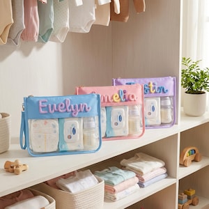 Puede incluir: Tres estuches rectangulares de plástico transparente con bordes de tela de colores y cremalleras. Cada estuche presenta un nombre personalizado en letras de hilo texturizadas y coloridas. Contienen pañales, toallitas y biberones.