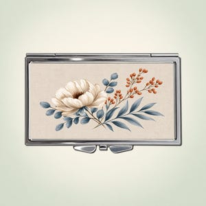 Peut inclure: Un étui rectangulaire en métal avec une finition argentée polie, orné d'une illustration centrale d'une grande fleur crème, de feuilles bleu-gris et de petites baies orange sur un fond beige texturé façon tissu avec un fermoir.
