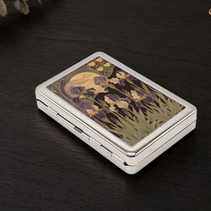 May include: A rectangular silver-toned metal case featuring a decorative top panel with a printed illustration of purple and cream-colored iris flowers, green foliage, and a large pale moon against a dark background, with a hinged side closure.