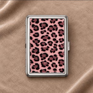 May include: A rectangular metal cigarette case featuring a pink and black leopard print pattern on the front panel. The case has a polished silver-tone frame with a hinged side and a secure latch mechanism for opening and closing the container.