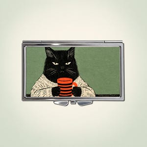 Pode incluir: Um estojo retangular de metal prateado com uma ilustração gráfica de um gato preto vestindo um suéter creme e segurando uma caneca listrada de laranja e preto contra um fundo verde-sálvia sólido. O estojo possui tampa e fecho.