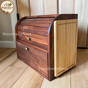 May include: A dark brown wooden bread box with a roll-top lid and a lower drawer, each featuring a small round brass knob. A light-colored wooden cutting board is attached to the right side of the unit. The text WoodvelleNest is visible.