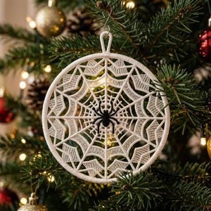May include: A circular white embroidered ornament featuring a detailed spider web pattern with a small black spider at the center. The ornament is constructed with a rigid frame and includes a small loop at the top for hanging on a tree branch.