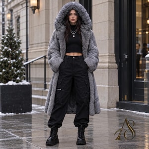 May include: A long, grey, textured faux fur coat with a large hood, worn over a black mock-neck crop top and black cargo pants with side pockets. The outfit is completed with black platform boots. A gold logo is visible in the bottom right corner.