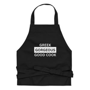 Op de afbeelding: Een zwart schort met een nekband en taillebanden. In het midden van het schort staat de witte tekst GREEK GORGEOUS GOOD COOK, waarbij het woord GORGEOUS is gemarkeerd in een wit rechthoekig kader, wat een duidelijk contrast vormt met de stof.
