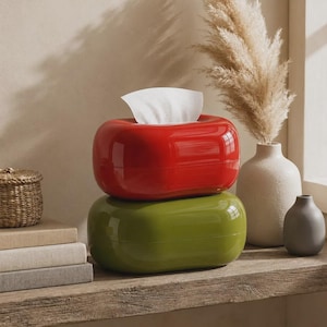 May include: Two stacked oval-shaped tissue boxes with a glossy finish. The top box is bright red and the bottom box is olive green. A single white tissue is pulled through the top opening of the red box, extending upward from the center.