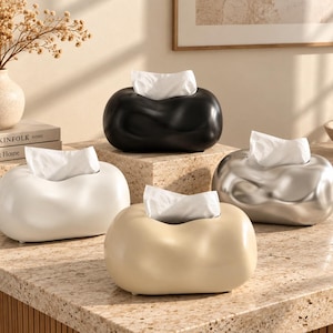 Puede incluir: Cuatro cajas de pañuelos ovaladas con una textura de superficie irregular, orgánica y lisa. El conjunto incluye una caja negra, una blanca, una beige y una plateada metálica, cada una con una abertura central y pañuelos blancos.