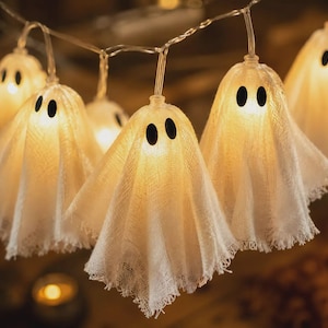 May include: A string of LED fairy lights featuring multiple white fabric ghost covers. Each ghost is constructed from a textured, semi-sheer white cloth with frayed edges and two small, solid black oval shapes serving as eyes, illuminated from within.