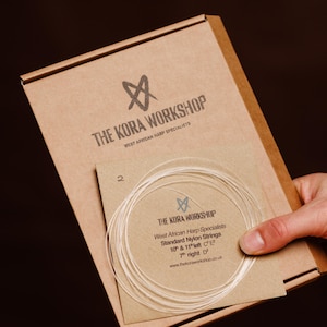 May include: A brown cardboard box labeled The Kora Workshop West African Harp Specialists containing a smaller envelope with coiled clear nylon strings. The envelope text reads Standard Nylon Strings 10th & 11th left C2, 7th right D2.