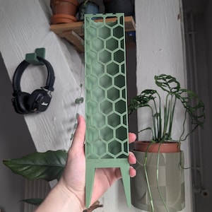 Op de afbeelding: Een saliegroene plantensteun met een herhalend zeshoekig honingraatpatroon. De structuur is rechthoekig en heeft twee puntige uiteinden aan de onderkant om in de aarde te steken, bedoeld als verticale ondersteuning voor klimmende kamerplanten.