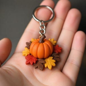 Op de afbeelding: Een sleutelhanger met een centrale oranje pompoen omringd door een krans van kleine, veelkleurige herfstbladeren in tinten geel, rood en bruin. De pompoen en bladeren zijn met een korte ketting bevestigd aan een zilverkleurige metalen ring.