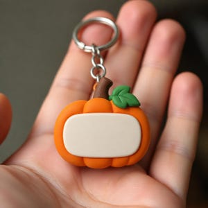 Op de afbeelding: Een oranje pompoenvormige sleutelhanger met een bruine steel, een enkel groen blaadje en een glad, leeg, lichtbeige rechthoekig vlak aan de voorzijde voor personalisatie, bevestigd aan een zilverkleurige metalen ring met een korte ketting.