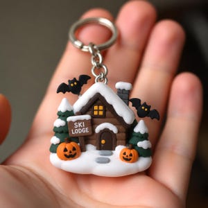 May include: A keychain featuring a miniature brown log cabin with a snow-covered roof, two green pine trees, and two orange jack-o'-lanterns. The cabin has a sign that reads SKI LODGE and two black bats with yellow eyes flying above it.