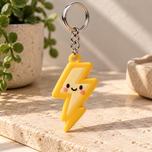 May include: A yellow lightning bolt keychain featuring a smiling face with black eyes, a small black mouth, and pink circular cheeks. The charm is attached to a silver-toned metal split ring by a short chain, creating a simple and functional accessory.