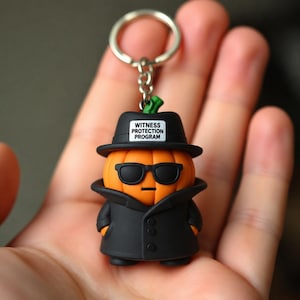 Może przedstawiać: Brelok przedstawiający pomarańczową dynię w czarnym kapeluszu typu fedora z białą etykietą z napisem WITNESS PROTECTION PROGRAM, czarnych okularach przeciwsłonecznych i czarnym płaszczu, przymocowany do srebrnego metalowego kółka i łańcuszka.