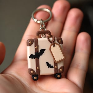 May include: A cream-colored suitcase keychain featuring brown straps, dark brown corner accents, and two black bat silhouettes. The luggage-shaped charm includes a small, blank, light brown rectangular luggage tag and a silver metal keyring.