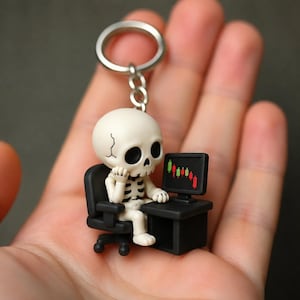 Può includere: Un portachiavi con una piccola statuetta di scheletro color bianco sporco seduta su una sedia da ufficio nera davanti a una scrivania nera. Lo scheletro appoggia il mento su una mano davanti a un monitor che mostra un grafico azionario.