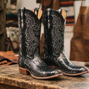 May include: A pair of black cowboy boots featuring a textured snakeskin-patterned foot and a tall shaft with intricate grey scrollwork embroidery. The boots have a pointed toe, a stacked wooden heel, and silver-toned hardware accents at the top.