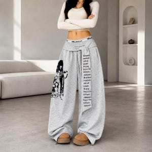 May include: High-waisted heather grey wide-leg sweatpants featuring a black graphic print on the left leg and a long vertical fabric strip on the front with printed black text. The waistband is elasticised with a white band showing black text.