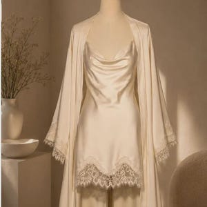 May include: A cream-colored satin slip dress with a cowl neckline and a matching robe featuring wide sleeves. Both pieces are adorned with intricate white lace trim along the hemline and sleeve cuffs, displayed on a mannequin stand.