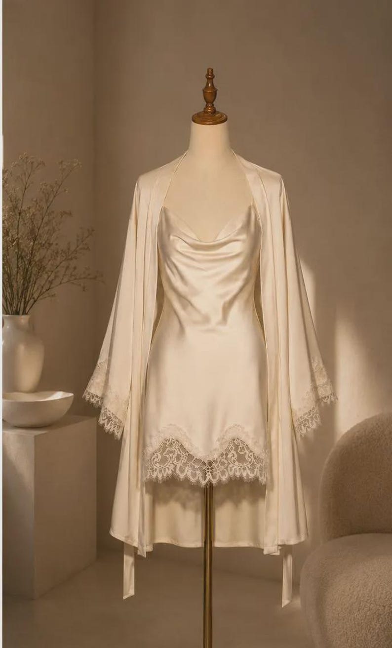 May include: A cream-colored satin slip dress with a cowl neckline and a matching robe featuring wide sleeves. Both pieces are adorned with intricate white lace trim along the hemline and sleeve cuffs, displayed on a mannequin stand.