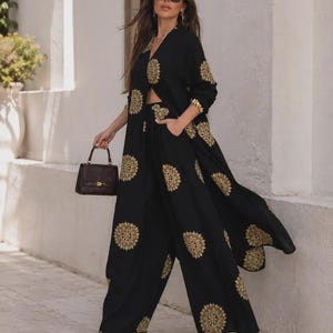 May include: A black two-piece set consisting of wide-leg trousers and a long-sleeved duster cardigan, both featuring a repeating gold circular mandala-style print. The outfit is paired with a small dark brown structured handbag and gold sandals.
