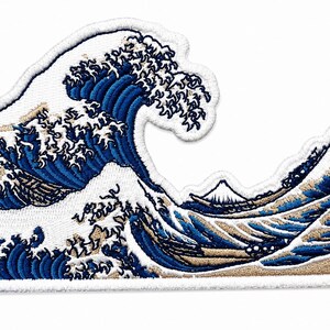 Puede incluir: Un parche bordado que presenta una representación estilizada de una gran ola oceánica rompiendo en tonos azul oscuro y blanco, con un pequeño pico de montaña visible al fondo sobre una base color canela, bordeado con costura blanca.