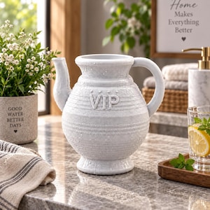 May include: A light gray ceramic pitcher with a speckled finish and a rounded body. The word VIP is embossed on the front, accompanied by horizontal grooved lines encircling the vessel. It features a curved handle and a short, tapered spout.