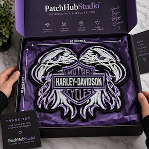 May include: A large embroidered Harley-Davidson patch featuring two eagle heads flanking a central shield logo with the text MOTOR HARLEY-DAVIDSON CYCLES. The patch measures 10 inches by 10 inches and is presented in a black box with purple tissue paper.