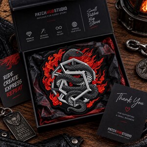 May include: A black embroidered patch featuring a grey snake coiled around a white geometric frame, surrounded by red flames. The patch is presented in a black box labeled PatchHubStudio with text including Ride Create Express Repeat.